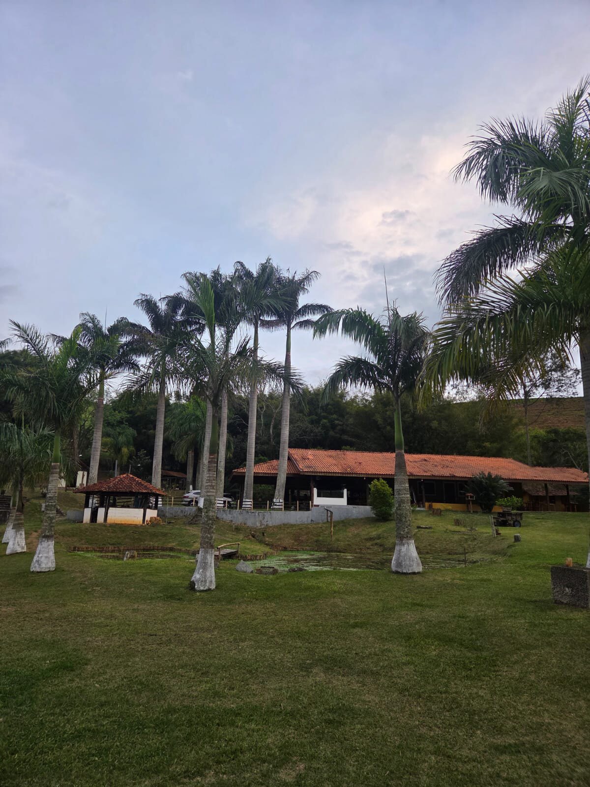 Gramado amplo com palmeiras, coreto de telhado de barro e o restaurante ao fundo